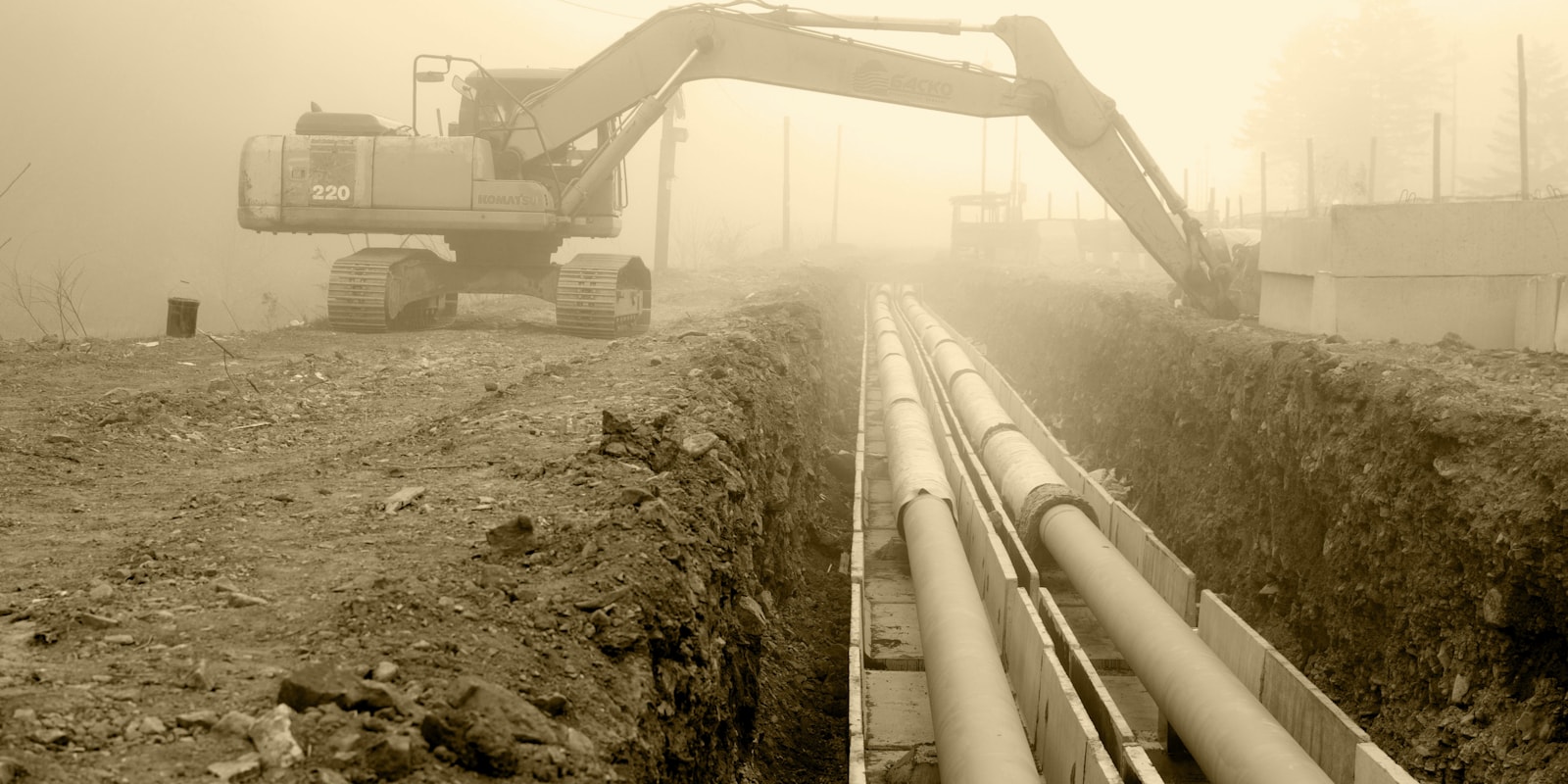 Tranchée de raccordement avec canalisations en cours de pose, pelleteuse en arrière-plan